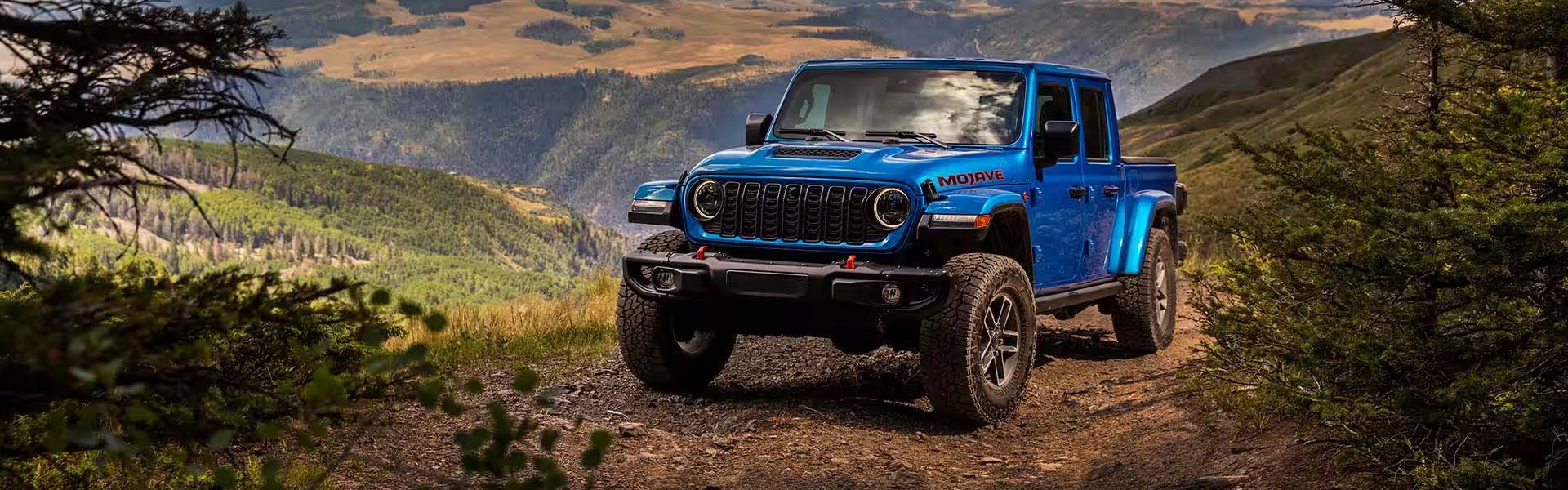 Blue Jeep Gladiator pickup truck on a dirt mountain trail overlooking a forested valley.