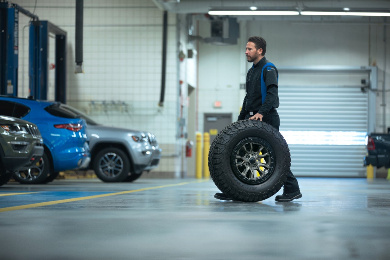Man in a garage uniform rolling a large off-road tire past parked cars in a clean service bay.